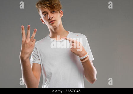 handsome emotional teenager boy posing in studio against gray, guy in a gray t-shirt shows the number three with his fingers Stock Photo