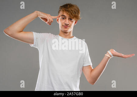 handsome emotional teenager boy posing in studio against gray, a guy in a gray T-shirt twists his finger at the temple, crazy Stock Photo