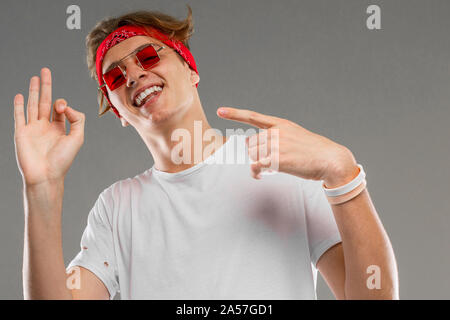 handsome emotional teenager boy posing in studio against gray, guy in a gray t-shirt and red sunglasses gestures ok Stock Photo