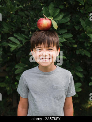 Happy young boy balancing an apple on his head outside by a tree. Stock Photo