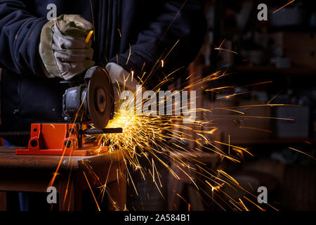 Hands of a craftsman with working gloves cutting an iron bar with the electric angle grinder which sprays many hot sparks in the dark workshop, danger Stock Photo