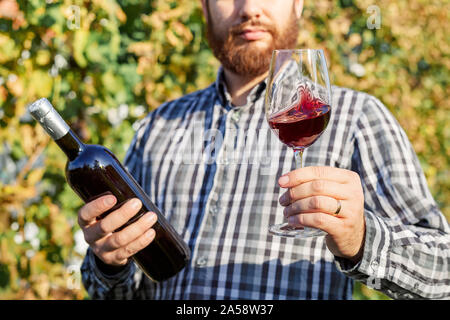 Handsome wine maker holding in his hand bottle and a glass of red wine and tasting it, checking wine quality while standing in vineyards. Small Stock Photo