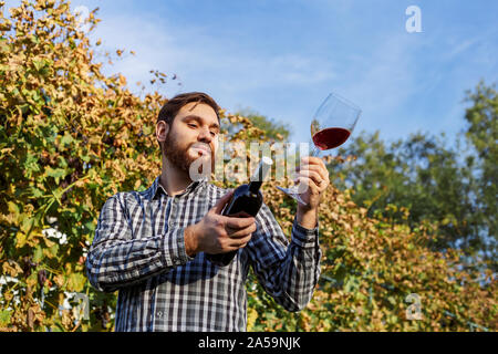 Portrait of handsome wine maker holding in his hand bottle and a glass of red wine and tasting it, checking wine quality while standing in vineyards Stock Photo