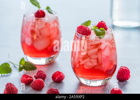 Two refreshing red translucent drinks with ice and raspberries. The raspberry juice is garnished with a green leaf, and there are pieces of ice and ra Stock Photo
