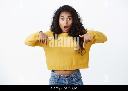 Female customer gasps from astonishment as pointing down. Emotional amazed african-american girl in yellow sweater drop jaw amused, indicating Stock Photo