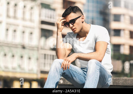 Outdoors Leisure. Stylish guy in sunglasses sitting on the street touching hair pensive Stock Photo