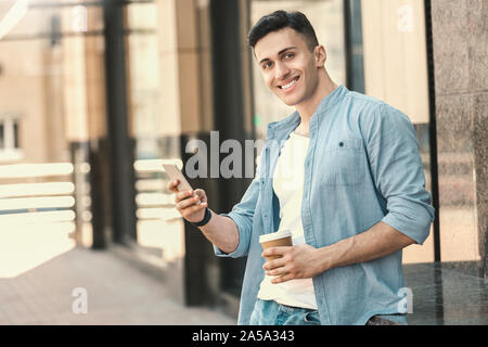 Outdoors Leisure. Stylish guy standing leaning back on the street with cup of coffee using app on smartphone smiling cheerful Stock Photo