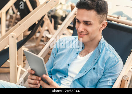 Outdoors Leisure. Stylish traveler sitting on the beach browsing digital tablet smiling joyful close-up Stock Photo
