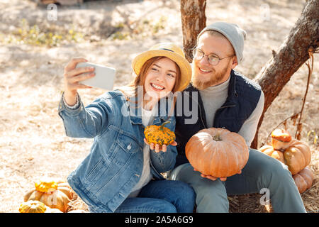 Halloween Preparaton Concept. Young couple sitting at garden with pumpkins taking selfie on phone posing smiling happy Stock Photo