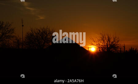 Black silhouette of a village house and trees at dark sunset. Round sun in orange sky in evening Stock Photo