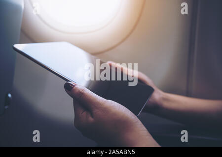 Closeup image of a woman holding and looking at black tablet pc next to an airplane window with clouds and sky background Stock Photo