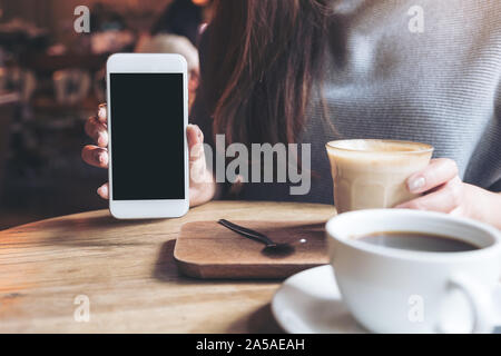 Mockup image of a woman holding and showing white mobile phone with blank black desktop screen with coffee cups on vintage wooden table Stock Photo