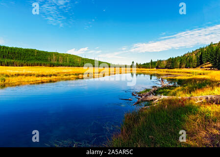 Peaceful blue river running through golden grassy valley with green forested mountains under a blue sky with white clouds. Stock Photo