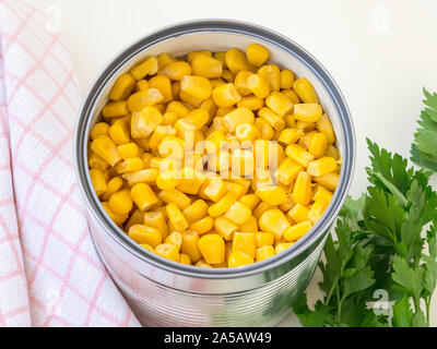 Canned sweet corn in a tin can and parsley on a table. Side dish and ingredient for salads. Vegetarian food. Top view. Stock Photo