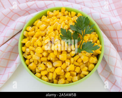 Canned sweet corn in a green bowl near checkered kitchen towel on a table. Side dish and ingredient for salads. Vegetarian food. Top view. Stock Photo