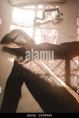 A black quilted winter jacket and retro camera lie on a bed illuminated by warm morning sunlight through window panes. Stock Photo