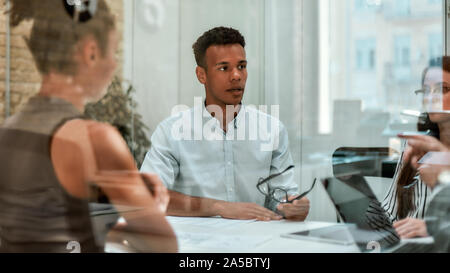 Business expert. Young afro american man holding eyeglasses and explaining something to his colleagues while sitting at the office table behind the Stock Photo