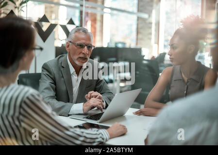 Brainstorming. Serious mature man in formal wear discussing something with his multicultural team while sitting at the office table Stock Photo