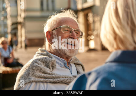 Happy senior bearded man in glasses looking at his wife and smiling while sitting on the bench together Stock Photo