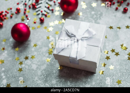 Silver gift with bow on grey snowy background with red Christmas balls and gold confetti stars. Stock Photo