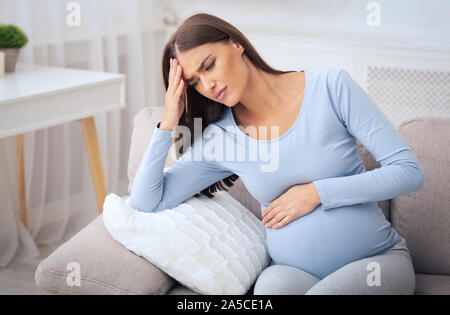 Pregnant Millennial Girl Having Headache Sitting On Sofa At Home Stock Photo