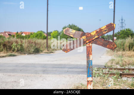 Level crossing sign, called crossbuck, saltire or Saint Andrews cross, rusted, standing on a countryside road which crosses and abandoned railway trac Stock Photo