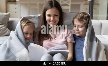 Happy mother with children covered with warm blanket reading book Stock Photo