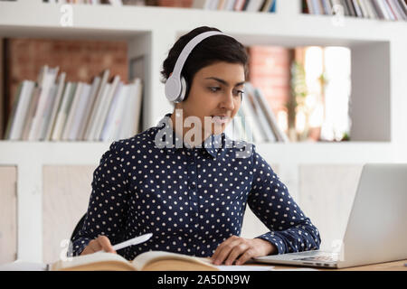 Indian girl in headphones do assignment in library using computer Stock Photo