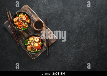 Stir fry with soba noodles, shrimps (prawns) and vegetables. Asian healthy food, stir fried meal in bowls over black background, copy space. Stock Photo