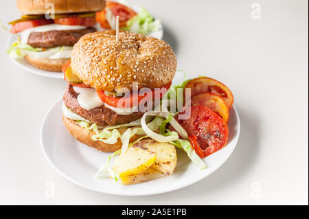 Horizontal shot two homemade hamburgers with a juicy meat cutlet, parmesan cheese, green salad, tomatoes and fried potatoes on a light background Stock Photo