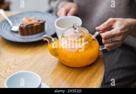 Girl pours a herbal berry tea from glass teapot into cap on wooden table background. Cafe in the street. Stock Photo