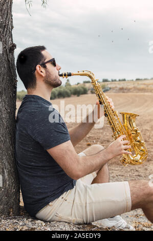 Young man sitting under a tree playing a saxophone on the countryside. Musician in a natural place with cloudy sky and birds on the background. Stock Photo