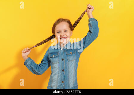 The child holds pigtails with his hands and laughs. Cheerful little girl in a denim shirt on a yellow background. Stock Photo