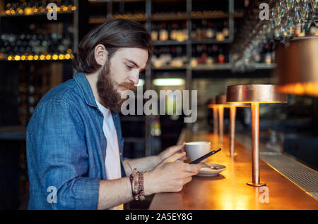 Young brown-haired bearded man having his coffee and using smartphone Stock Photo