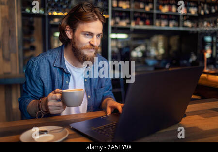 Young bearded freelancer checking email via laptop while having morning coffee in a loft bar Stock Photo