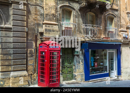 London-style iconic red phone box standing next to run-down old building and bright show-window of clothing store in Valletta, Malta. Stock Photo