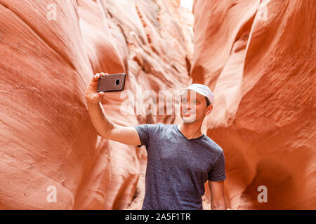 Orange red wave shape formations and man taking selfie picture with phone at narrow Antelope slot canyon in Arizona on footpath trail from Lake Powell