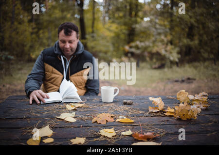 a young man sits in an autumn park at an old wooden table, drinks tea and reads a book. Warm dark autumn atmosphere, soft focus. In blur, copy space Stock Photo