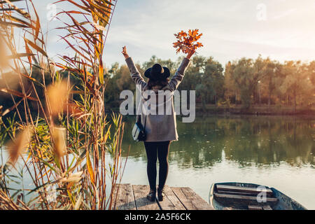 Happy woman walking on lake pier by boat raising hands and admiring autumn landscape. Fall season activities