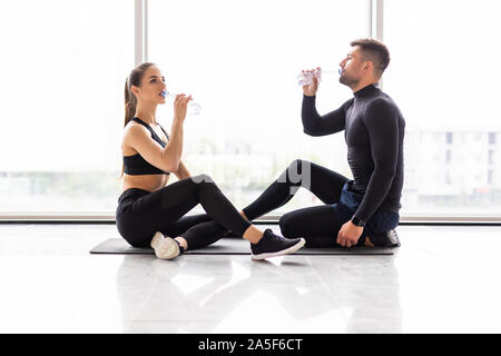 Tired fitness couple having rest and talking, drinking water at gym. Man and woman sitting on floor, break after training, healthy lifestyle concept Stock Photo