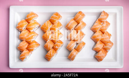 Philadelphia roll with salmon, cream cheese, rice and nori seaweed on a large rectangular white plate on a pink background. Top view Stock Photo