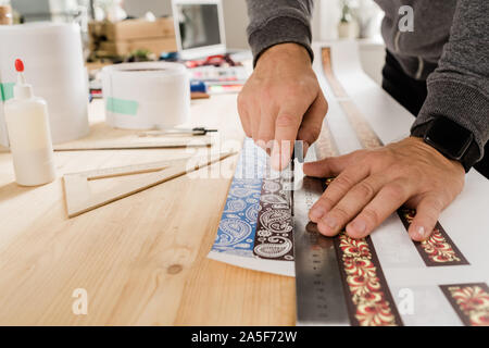 Hands of young craftsman or designer by wooden table using ruler and knife to cut ornamented paper to make collars Stock Photo