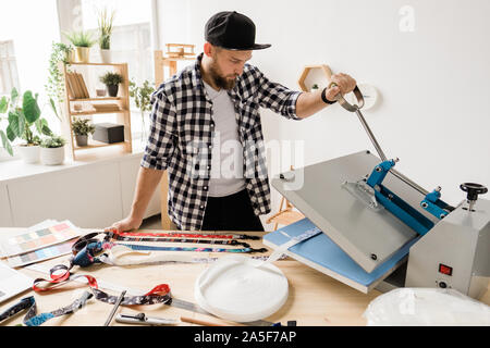 Young creative man printing or sticking decor on one of pet collar workpieces while using technical equipment Stock Photo
