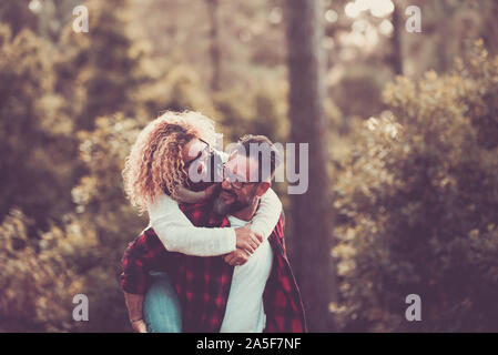 two adults having fun and laughing together at the wood with trees in the background - man holding her wife on his back - save the forest and change c Stock Photo