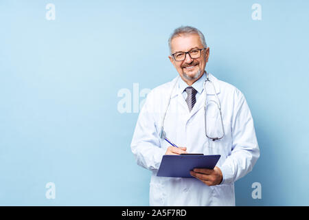 Smiling medical senior doctor with a stethoscope. On a blue background. The medic holds the folder in his hands and makes an appointment at the clinic Stock Photo