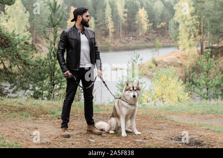 Young stylish man in black leather jacket and jeans standing on footpath in the forest or park while holding leash of his pet Stock Photo