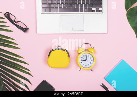 Office workplace with laptop computer, purse and alarm clock, with tropical decoration. Time is money concept for freelance on pink background. Stock Photo