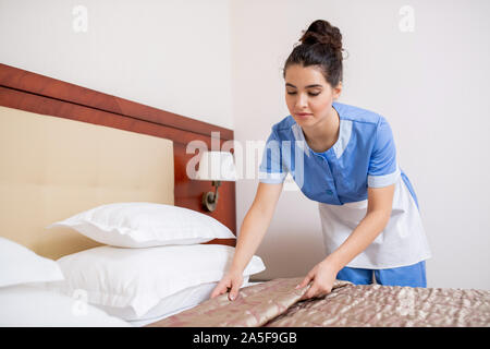 Pretty young brunette chamber maid in blue uniform making bed in hotel room while doing her work Stock Photo