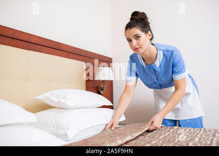Young brunette female in blue uniform of chamber maid looking at you while bending over bed in hotel room during work Stock Photo