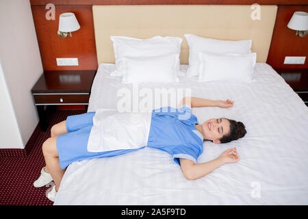 Happy young relaxed chamber maid in uniform lying on white clean bed while taking break on working day Stock Photo